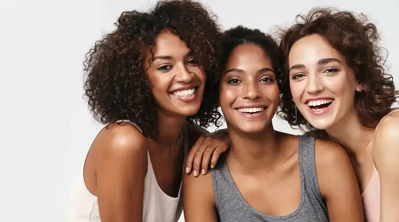 Three smiling women standing close together against a light background in a lifestyle beauty portrait